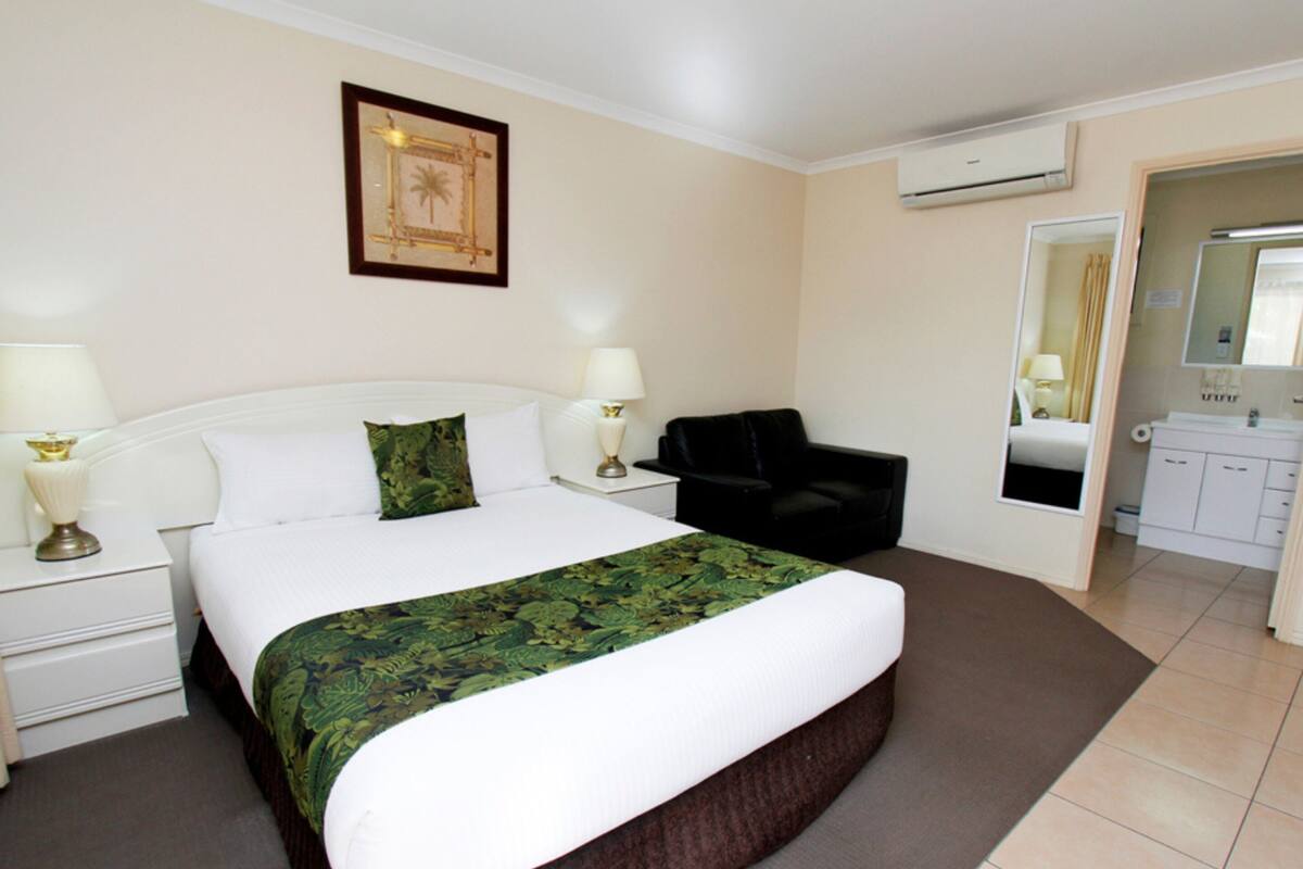 A hotel room with a bed, two bedside lamps, a black chair, a mirror, and a bathroom visible through an open door.