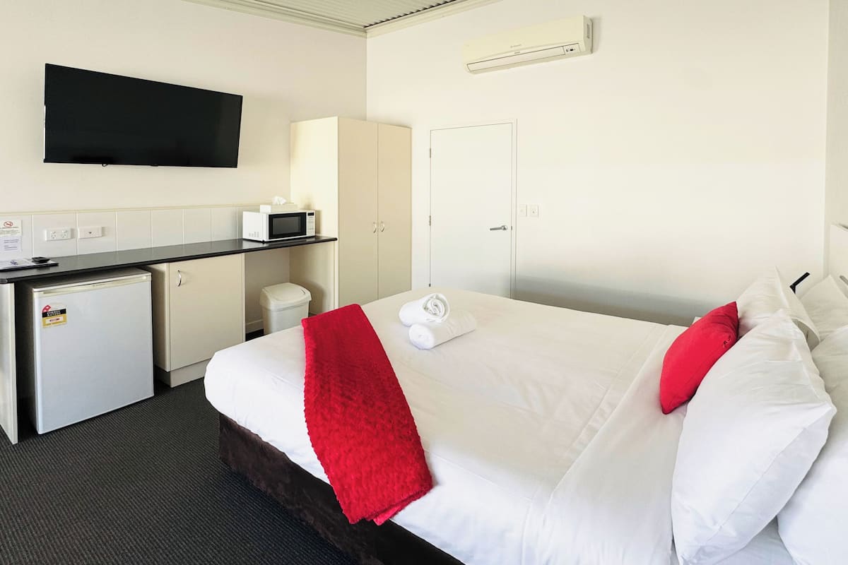 A hotel room with a bed, a desk with a microwave, a small refrigerator, a TV mounted on the wall, and a door.