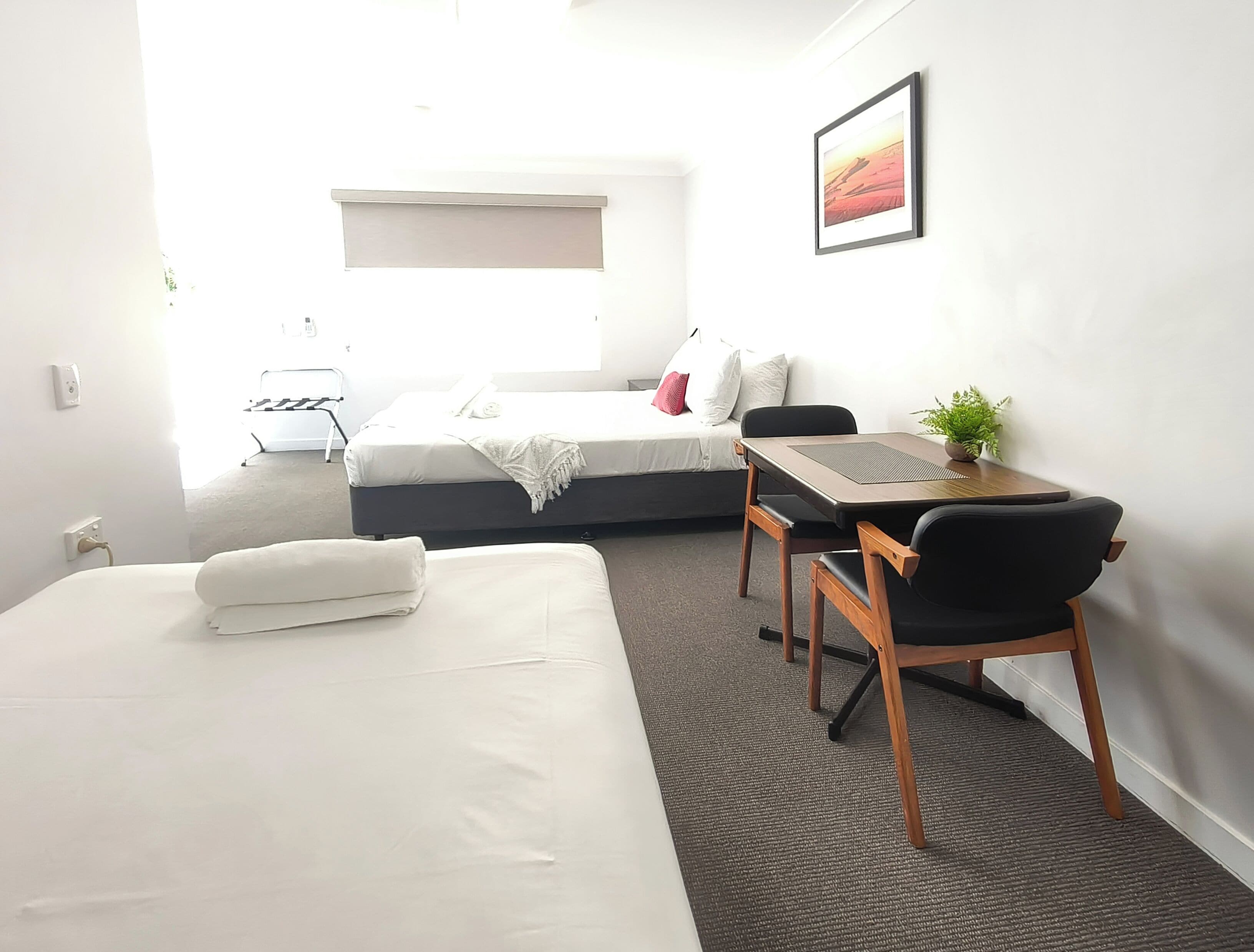 Habitación de hotel con dos camas, un escritorio con una silla, una mesita con una planta y un cuadro en la pared.