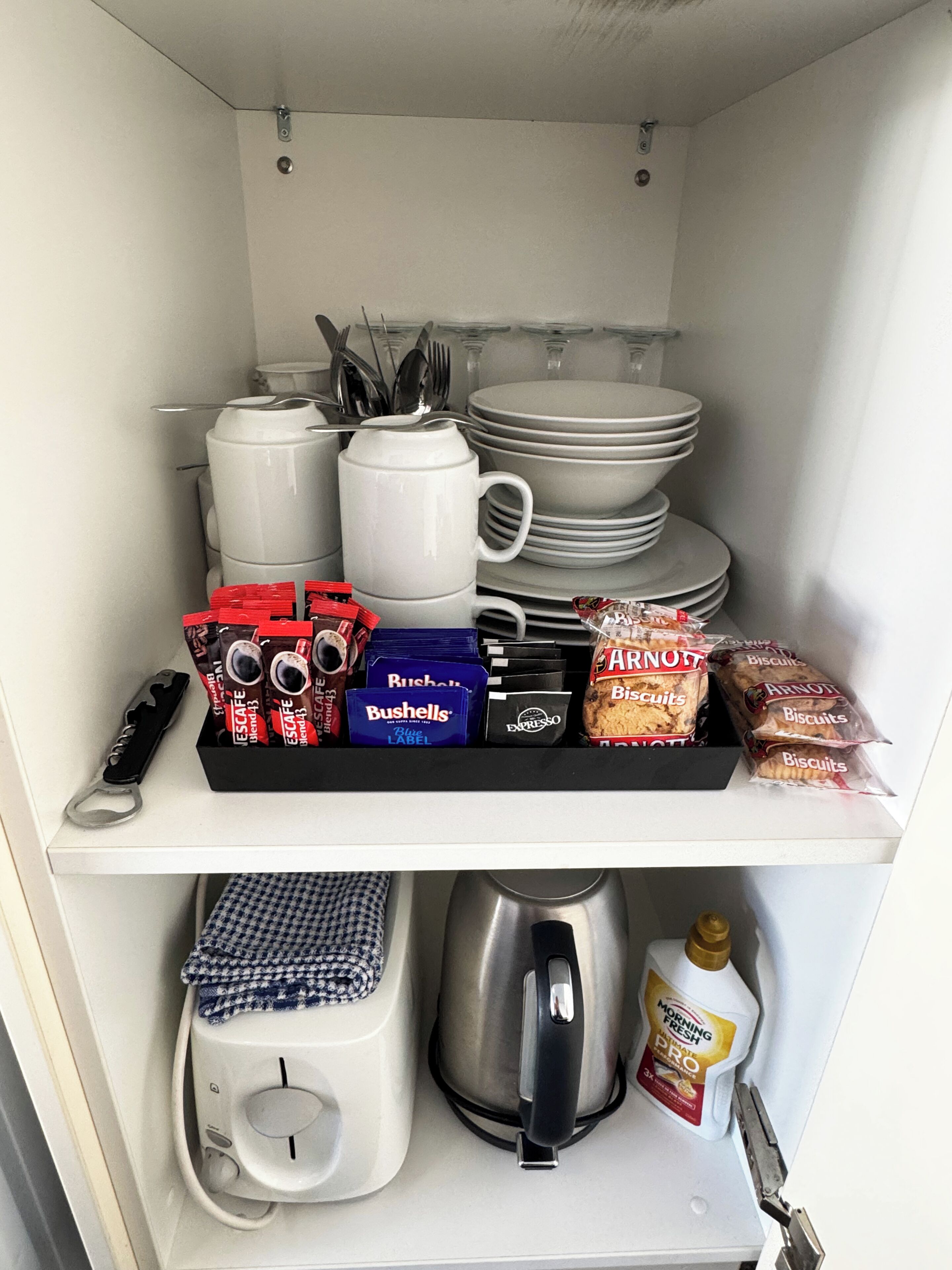 A kitchen pantry with storage shelves holding plates, mugs, utensils, coffee pods, biscuits, and a coffee maker.