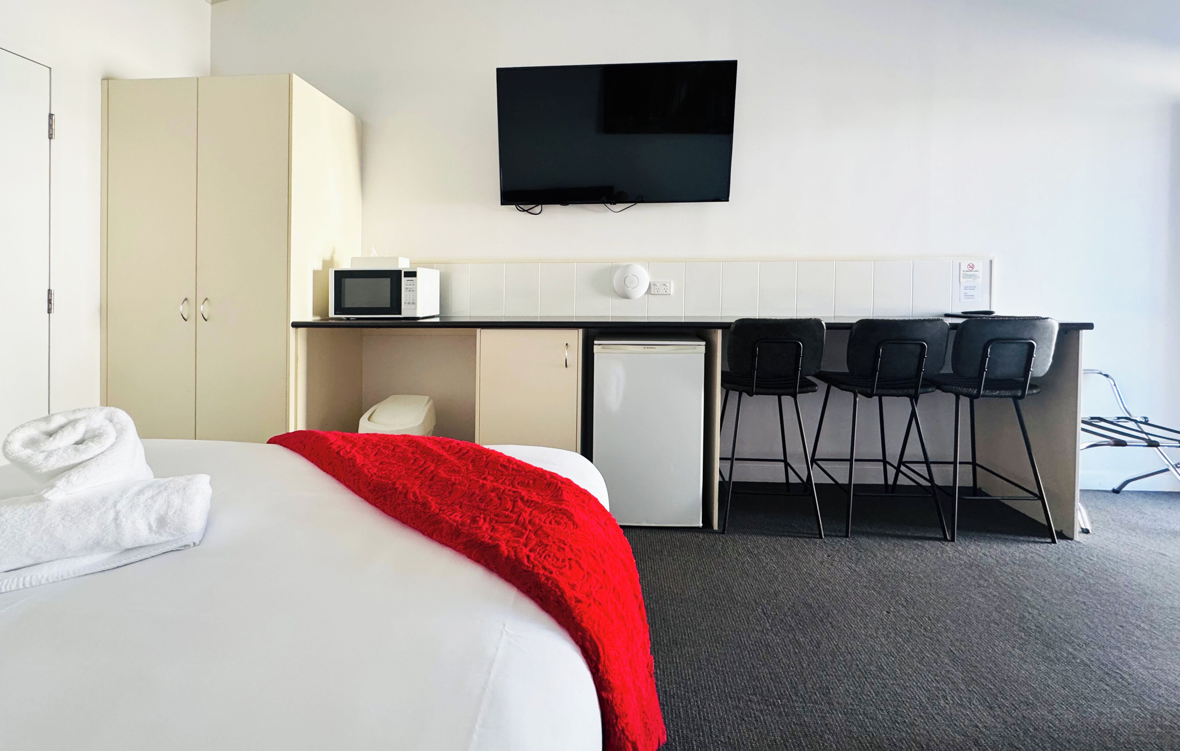 Habitación de hotel con cama, una manta roja, televisor montado en la pared, kitchenette con microondas y un refrigerador pequeño.
