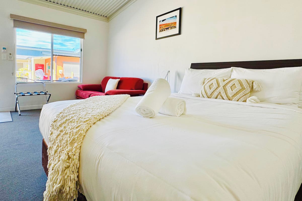 A hotel room with a large bed, a red chair, a window with a view, and a framed picture on the wall.