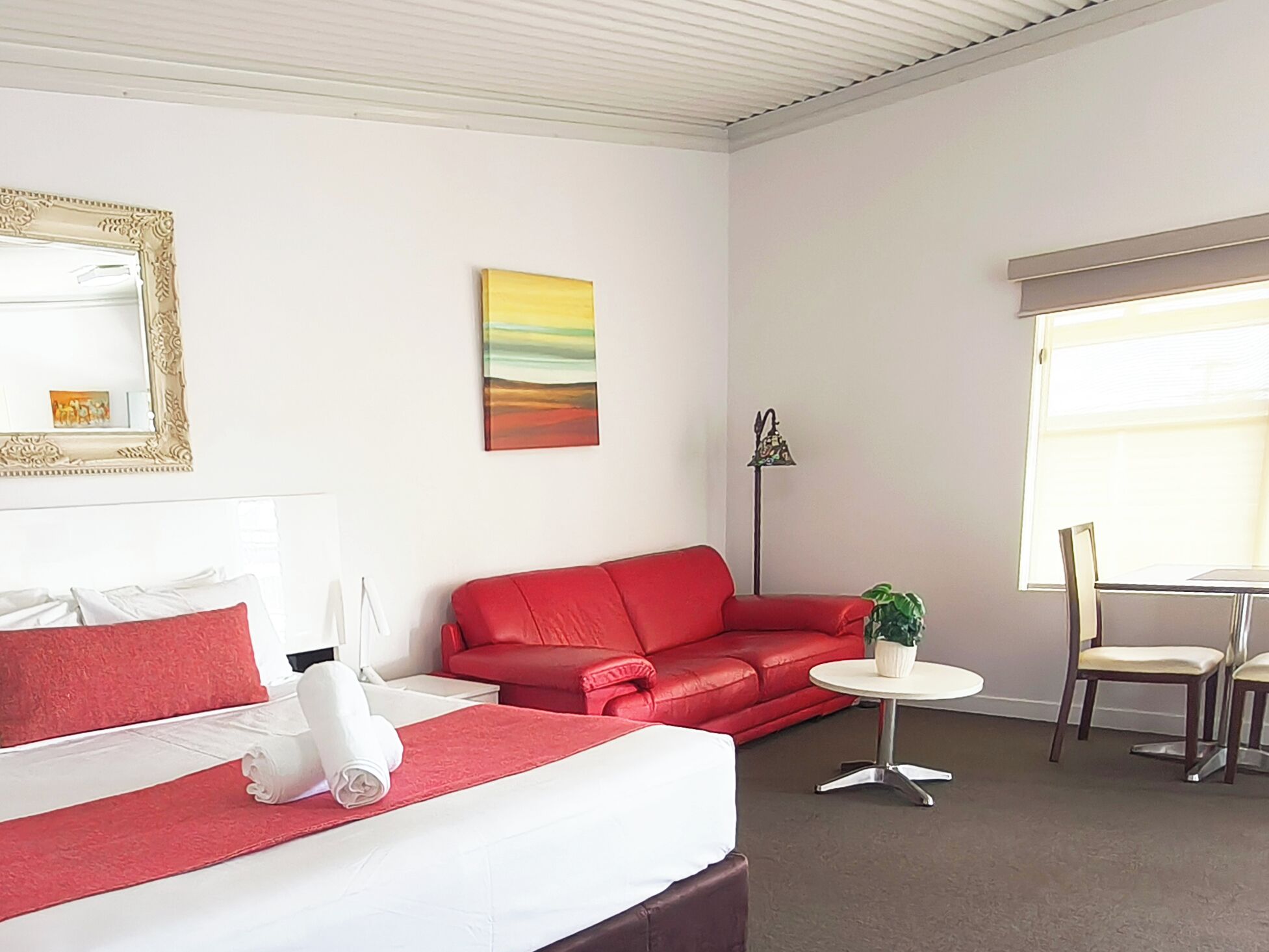 A hotel room with a bed, a red sofa, a small round table with a plant, a chair, and a window with blinds.