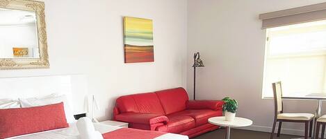 A hotel room with a bed, a red sofa, a small round table with a plant, a chair, and a window with blinds.