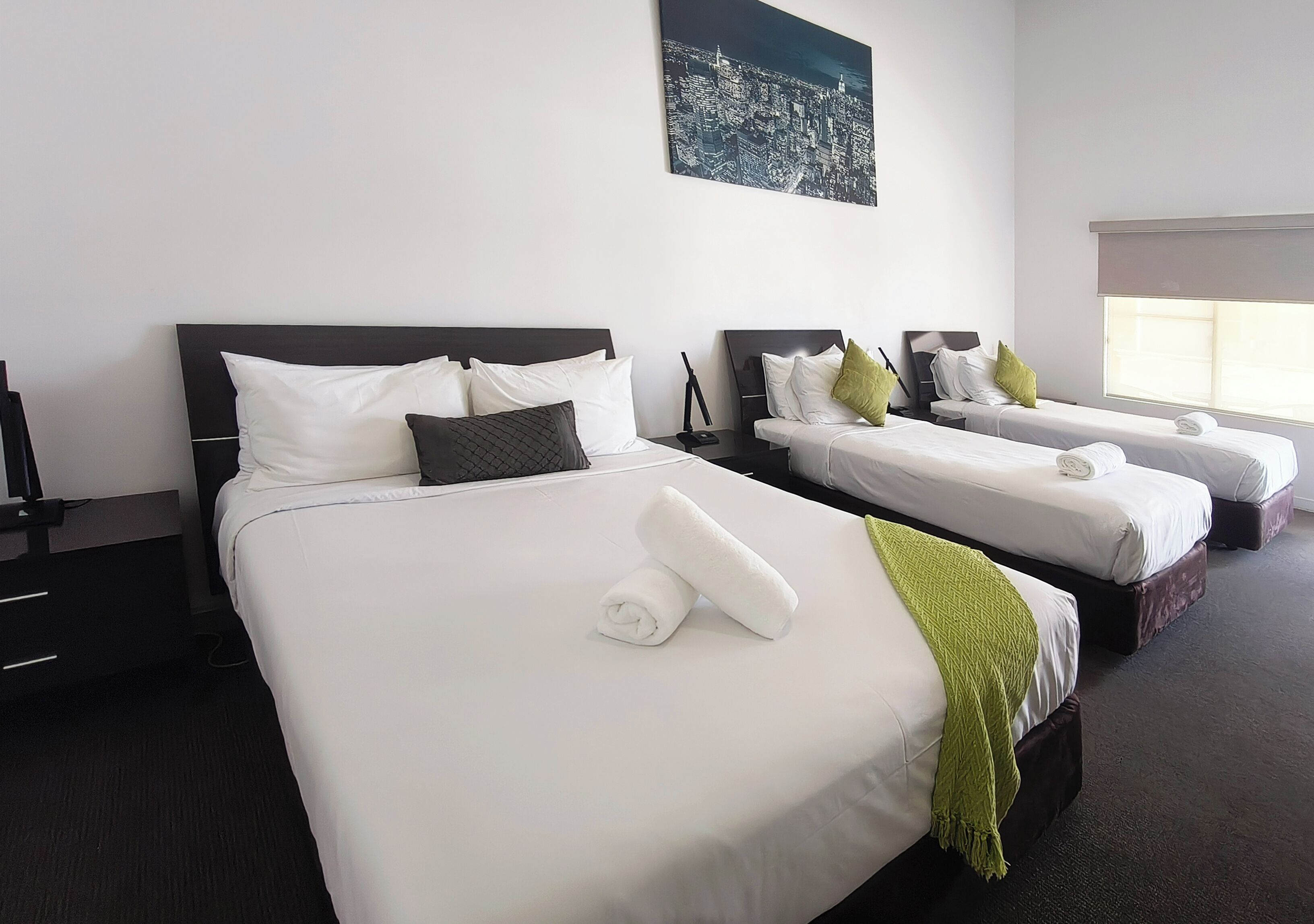 Ein Hotelzimmer mit drei Betten, jeweils mit weißer Bettwäsche und Kissen, einem Stadtbild an der Wand und einem großen Fenster, das für natürliches Licht sorgt.