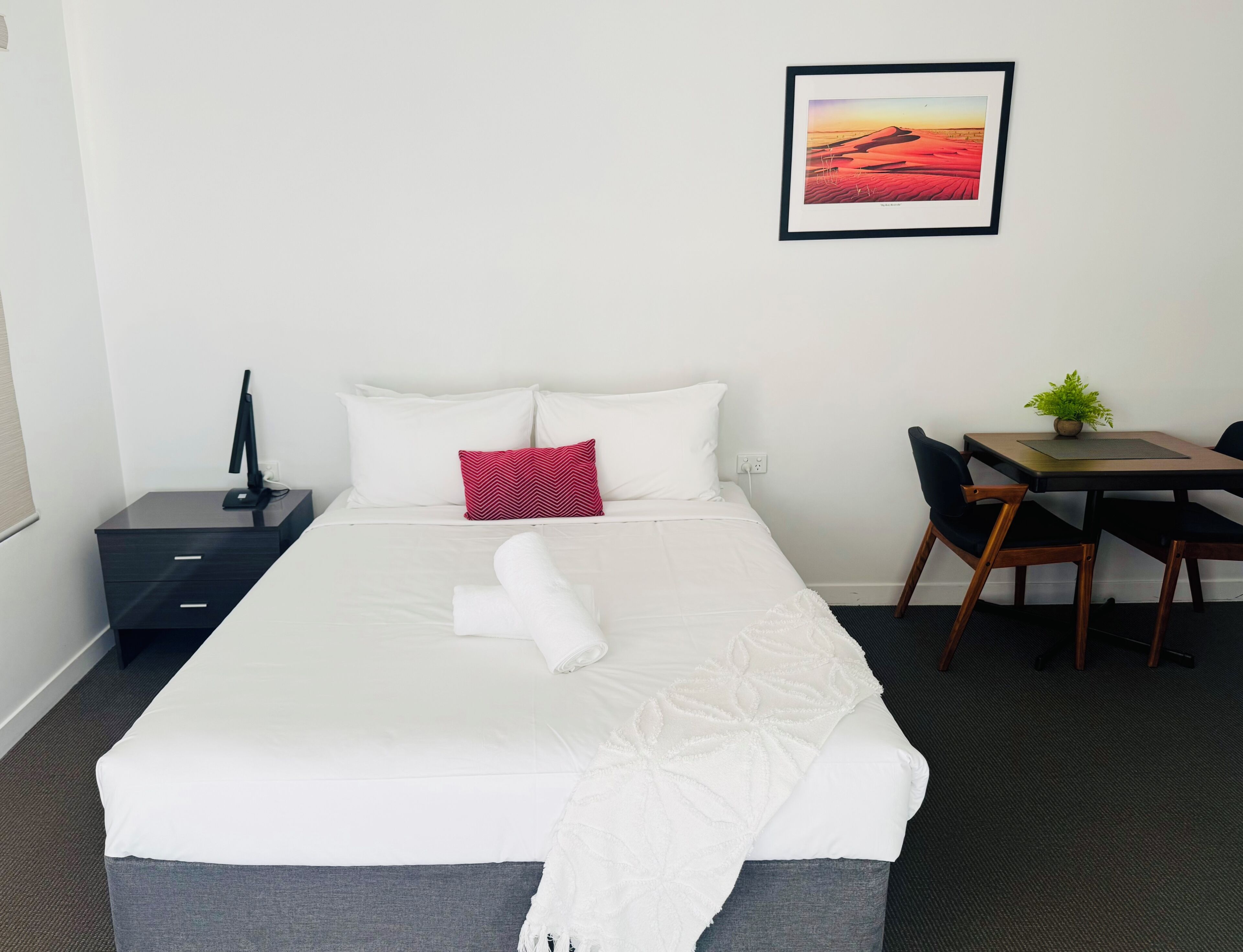 A neatly made bed with white linens, a red pillow, and a white towel. There is a small table with a plant, a chair, and a framed picture on the wall.
