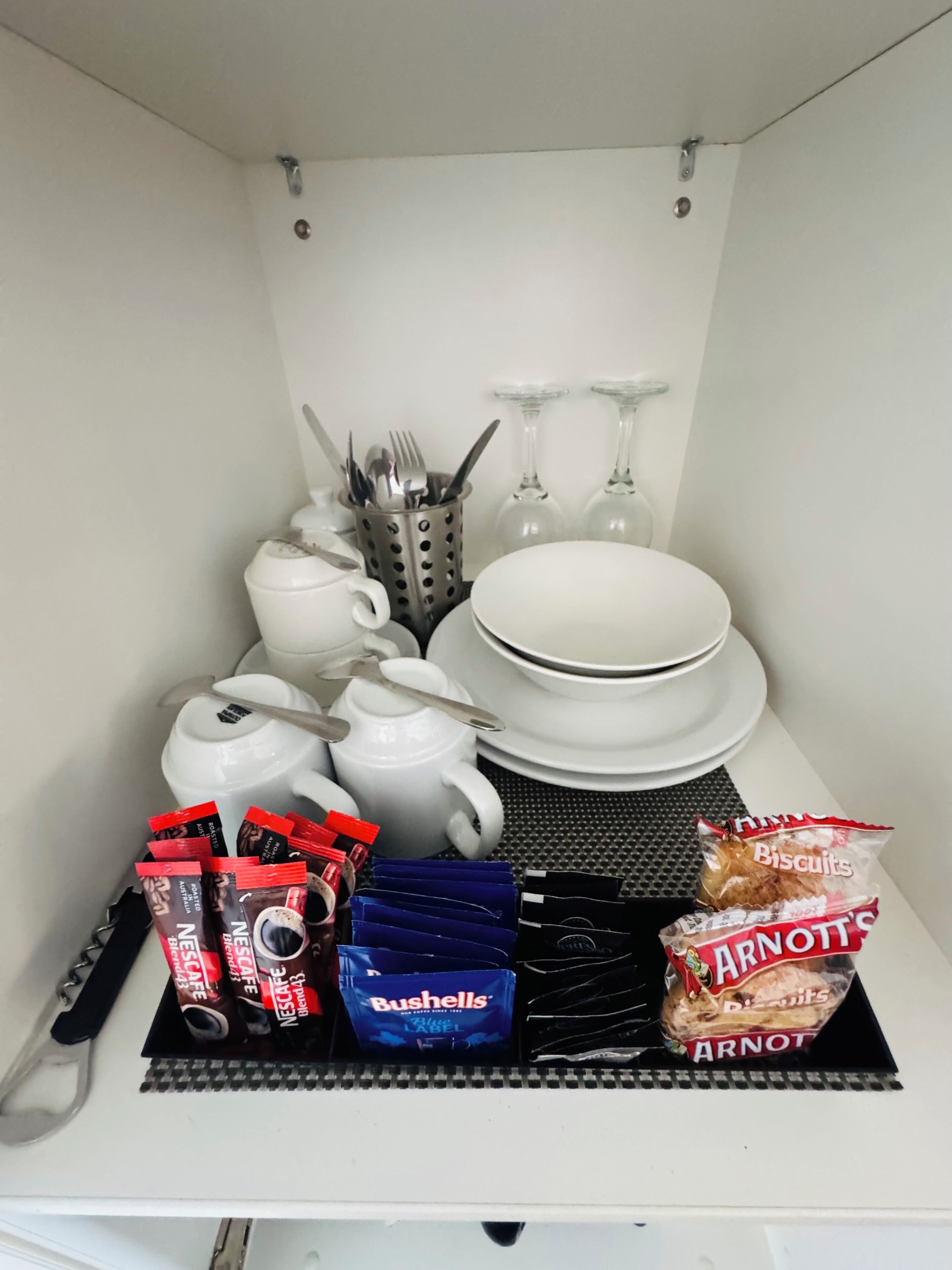 Una kitchenette con electrodomésticos blancos, un hervidor de agua, una cafetera, un tostador, un juego de platos, tazones, una copa de vino, un tenedor, una cuchara, un cuchillo, un bloque de cuchillos, un paquete de galletas, un paquete de café y un paquete de té.
