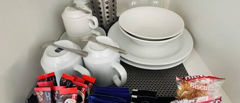 Una kitchenette con electrodomésticos blancos, un hervidor de agua, una cafetera, un tostador, un juego de platos, tazones, una copa de vino, un tenedor, una cuchara, un cuchillo, un bloque de cuchillos, un paquete de galletas, un paquete de café y un paquete de té.