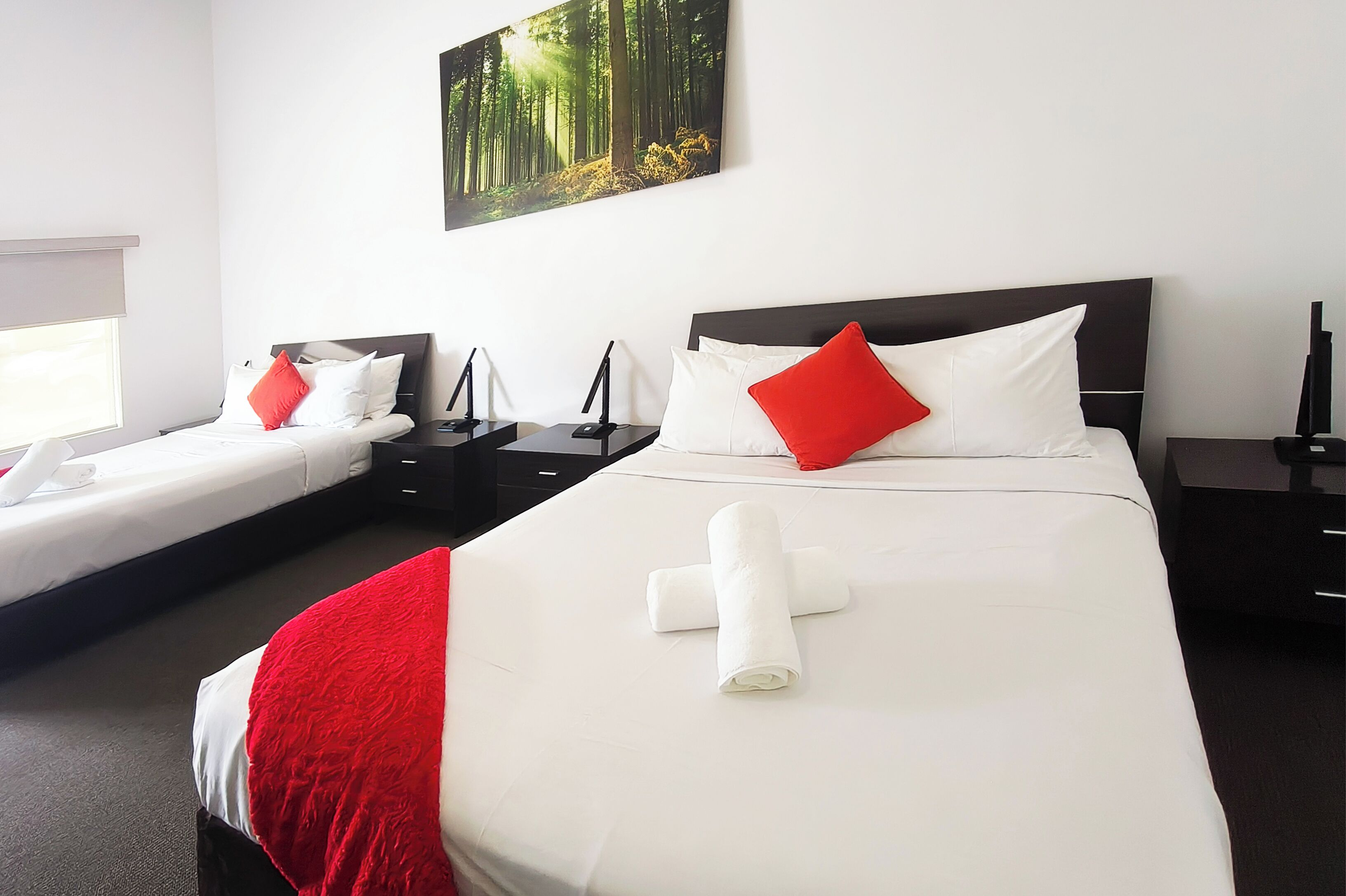 Une chambre d’hôtel avec deux lits, chacun agrémenté de draps blancs et d’oreillers rouges, une grande peinture représentant une forêt et un lit soigneusement fait avec une serviette roulée.