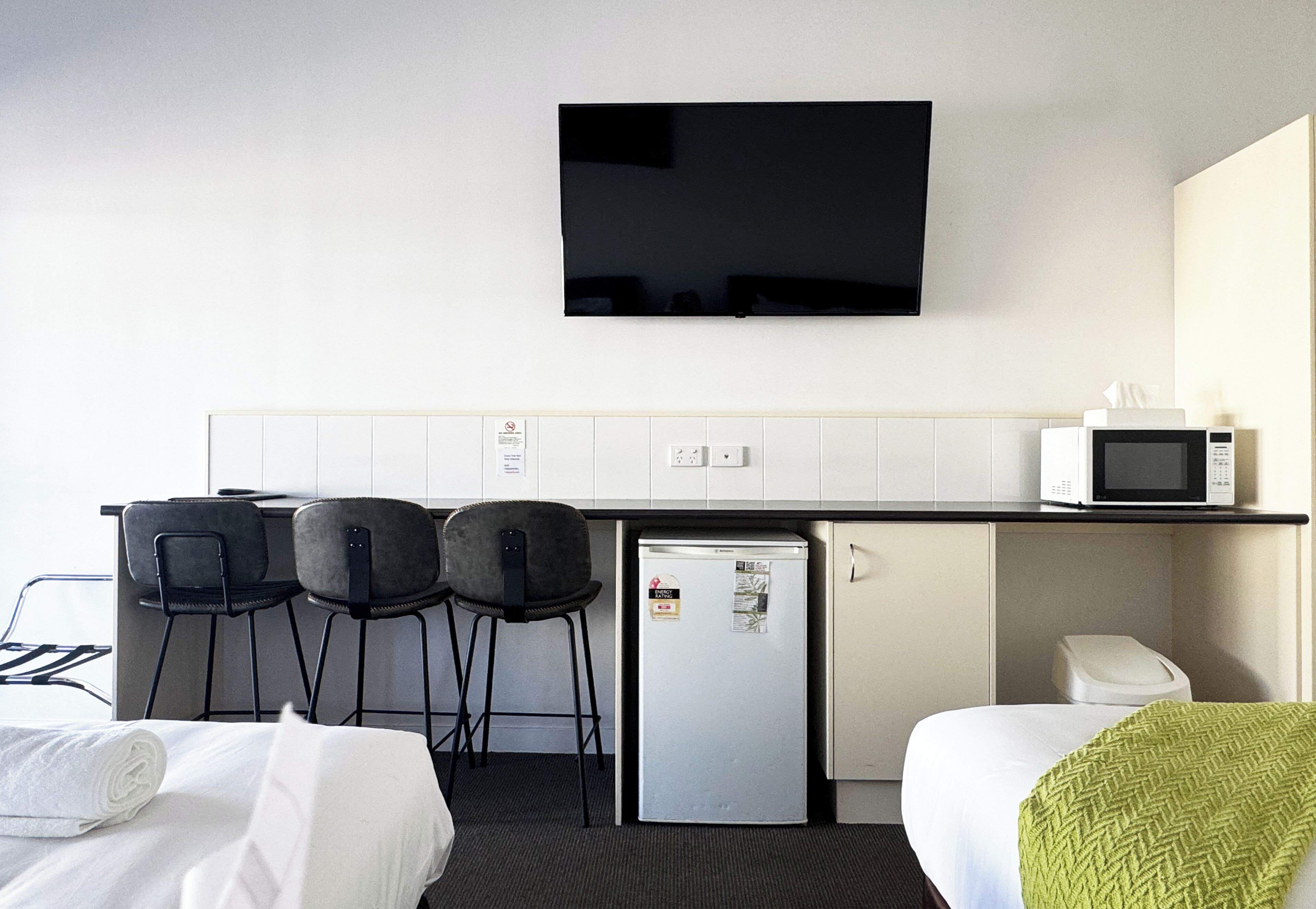 A compact hotel room with a kitchenette, a TV mounted on the wall, a small refrigerator, and two bar stools.