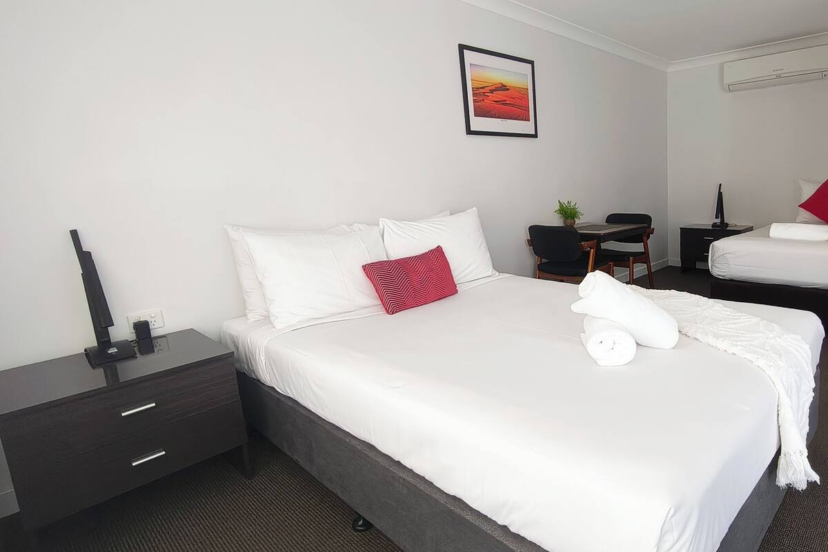 A hotel room with a bed, a desk, a chair, a small table, a plant, and a picture on the wall.