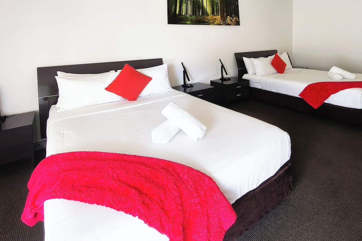 A hotel room with two beds, each with white linens, red pillows, and a red throw blanket. There is a painting on the wall, a nightstand with a lamp, and a door to the right.