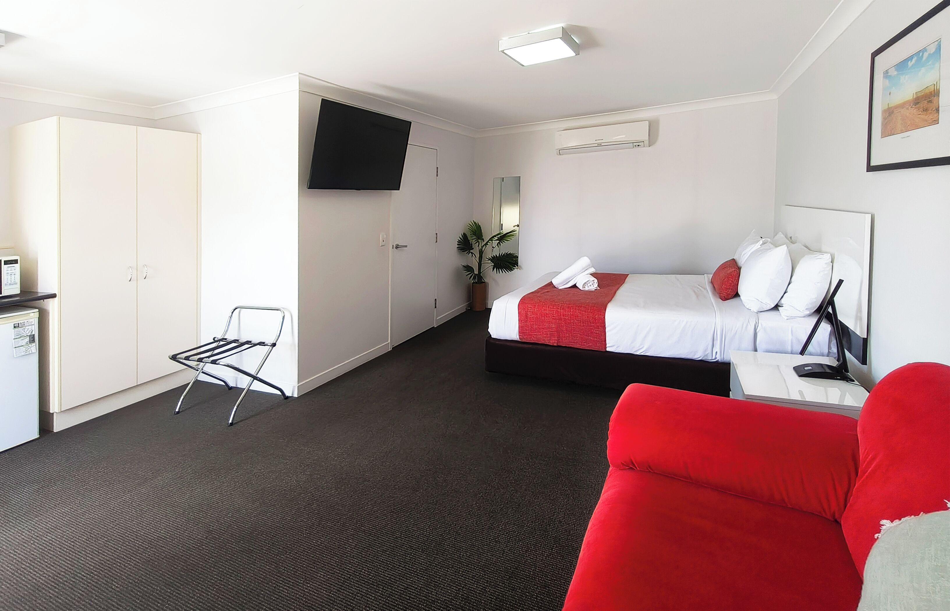 A hotel room with a bed, a red chair, a desk with a computer, a TV, and a wardrobe.