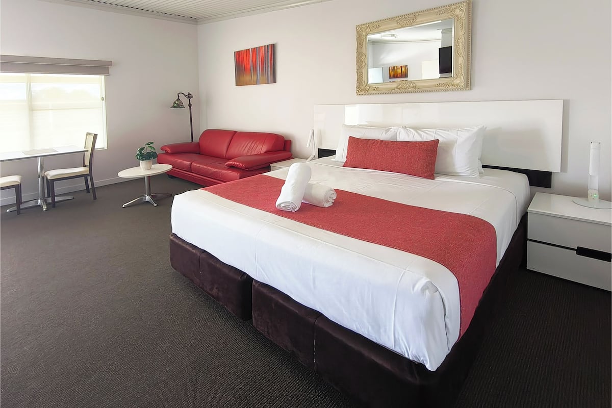 A hotel room with a large bed, a red sofa, a dining table, and a chair.