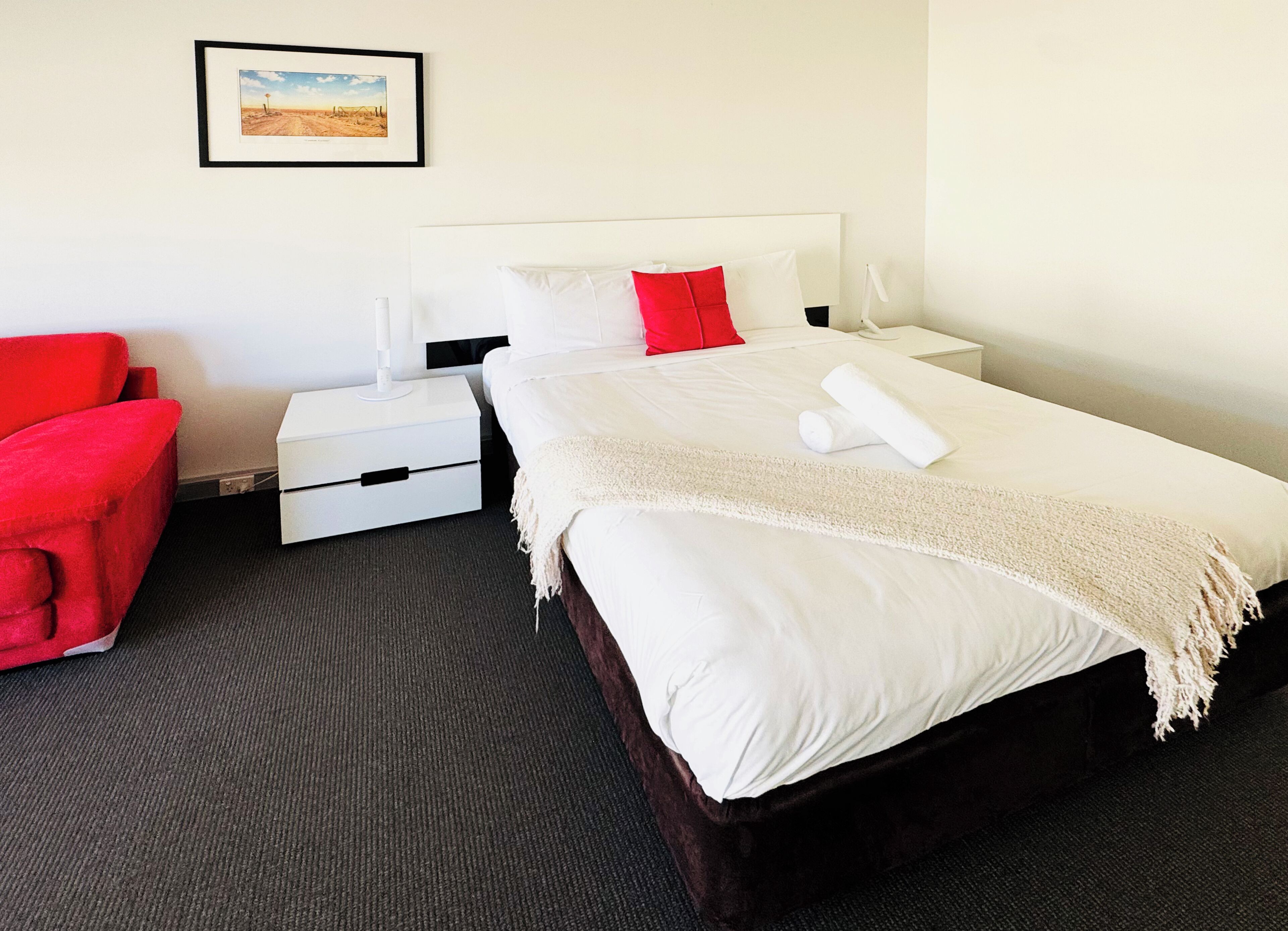 A hotel room with a bed, a nightstand, a red chair, a white bedside table, and a framed picture on the wall.