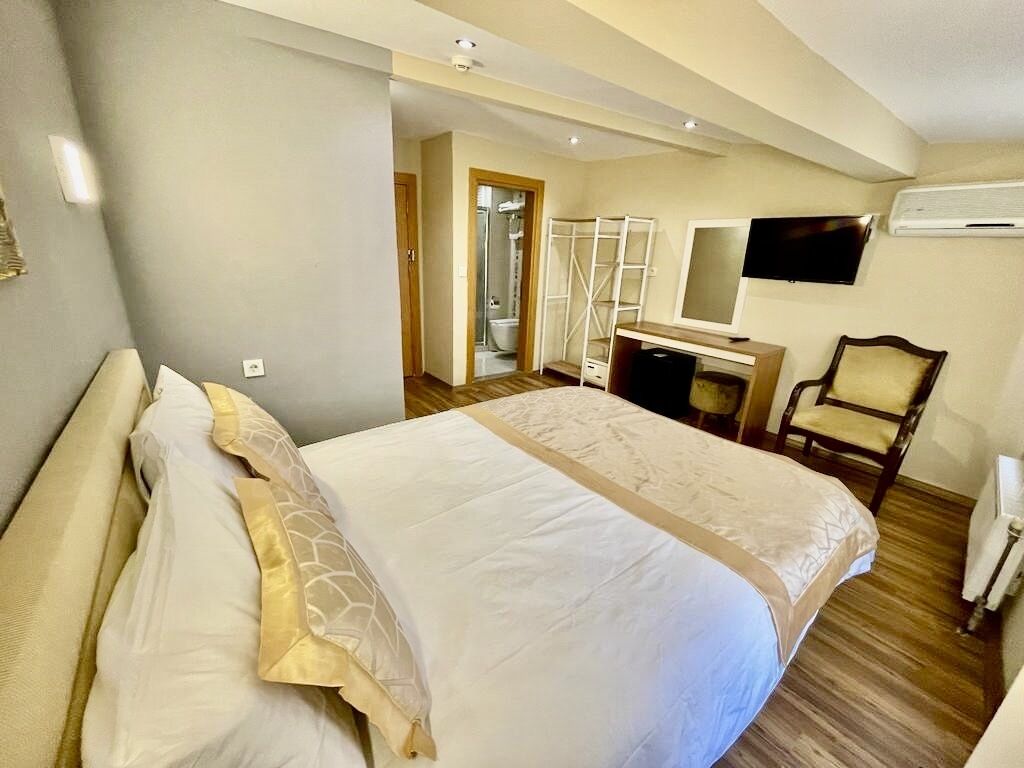 A hotel room with a large bed, a desk, a chair, a TV, and an air conditioning unit.