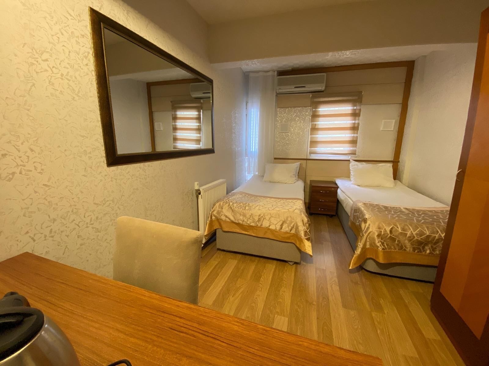 A compact hotel room with two single beds, a wooden desk, a chair, a large mirror, and an air conditioning unit.