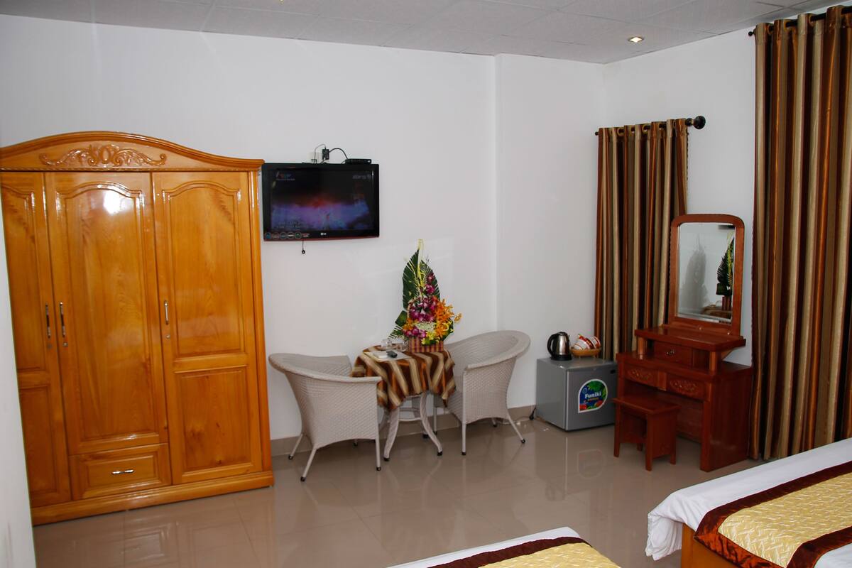 A hotel room with a wooden wardrobe, a television, a small table with a floral arrangement, a chair, a vanity with a mirror, and two beds.