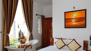 Une chambre d’hôtel comprenant un lit, un bureau avec une chaise, une fenêtre avec des rideaux, une armoire en bois et un tableau représentant un coucher de soleil.