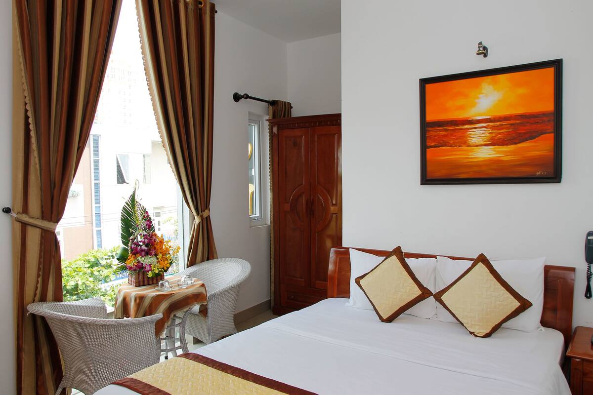 A hotel room with a bed, a desk with a chair, a window with curtains, a wooden wardrobe, and a painting of a sunset.