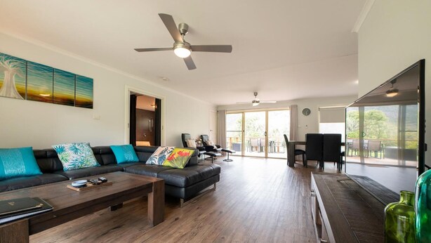 A spacious living area with a large sofa, a coffee table, and a dining table. There is a ceiling fan and a painting on the wall. - Halls Gap Valley Lodges (Halls Gap)