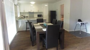 A modern kitchen with white cabinets, a microwave, and a refrigerator. There is a dining table with chairs and a small white table with a chair. - Halls Gap Valley Lodges (Halls Gap)