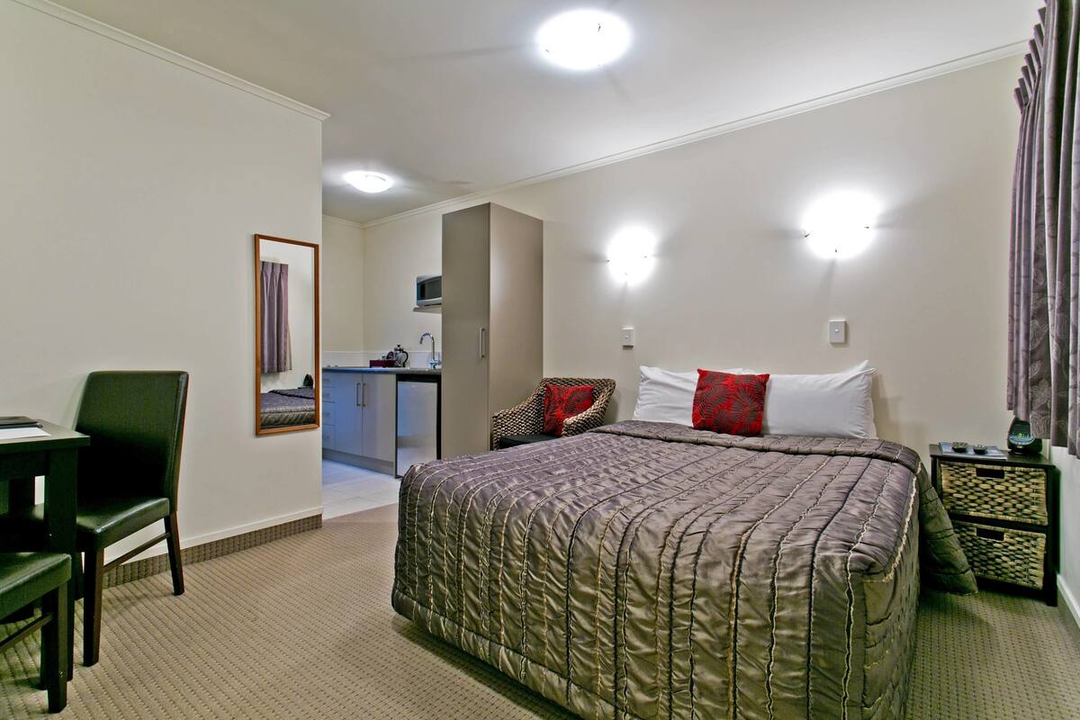 A hotel room with a large bed, a desk, a chair, and a small kitchenette in the background.