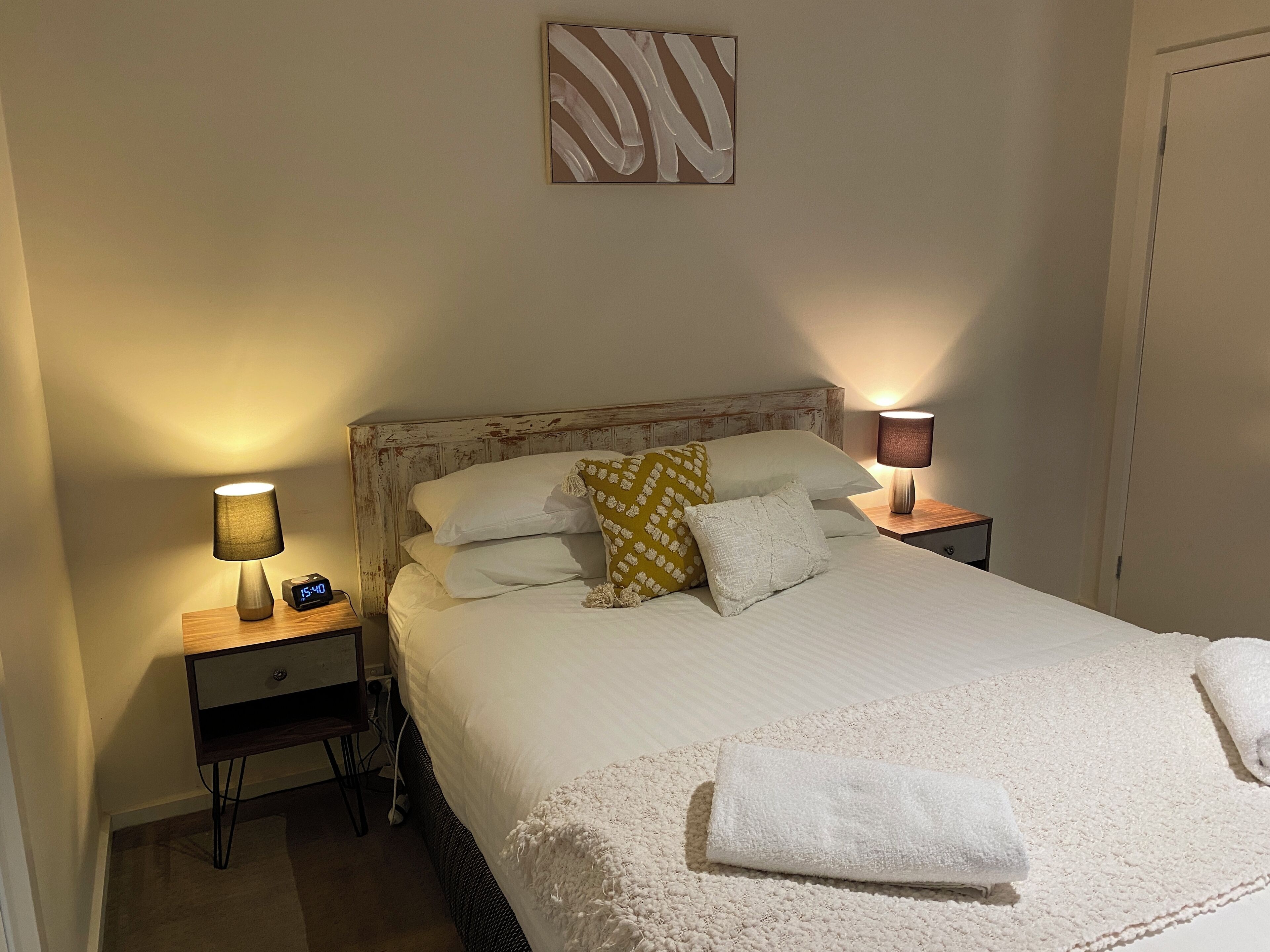 Een net opgemaakt bed met witte lakens, een geel gestreept kussen en een witte handdoek ernaast. Er staan twee nachtlampjes, waarvan er één brandt en de ander uit is. Een klein digitaal klokje is zichtbaar op het nachtkastje.