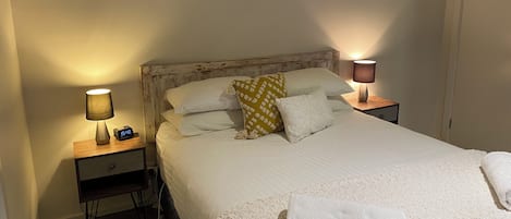 Een net opgemaakt bed met witte lakens, een geel gestreept kussen en een witte handdoek ernaast. Er staan twee nachtlampjes, waarvan er één brandt en de ander uit is. Een klein digitaal klokje is zichtbaar op het nachtkastje.