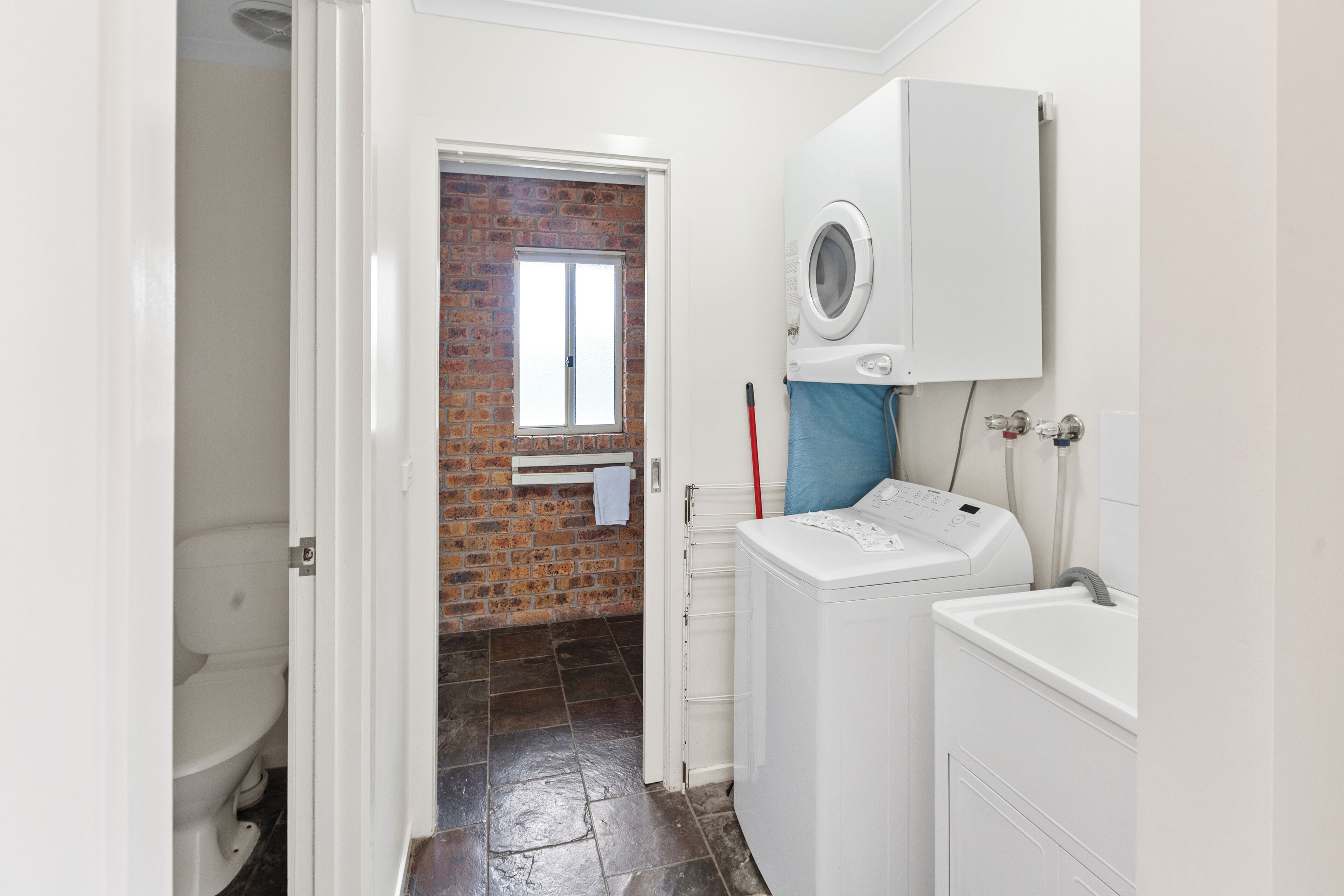 A laundry room with a washing machine, a dryer, and a sink. There is a door leading to a bathroom with a toilet and a window with a brick wall.