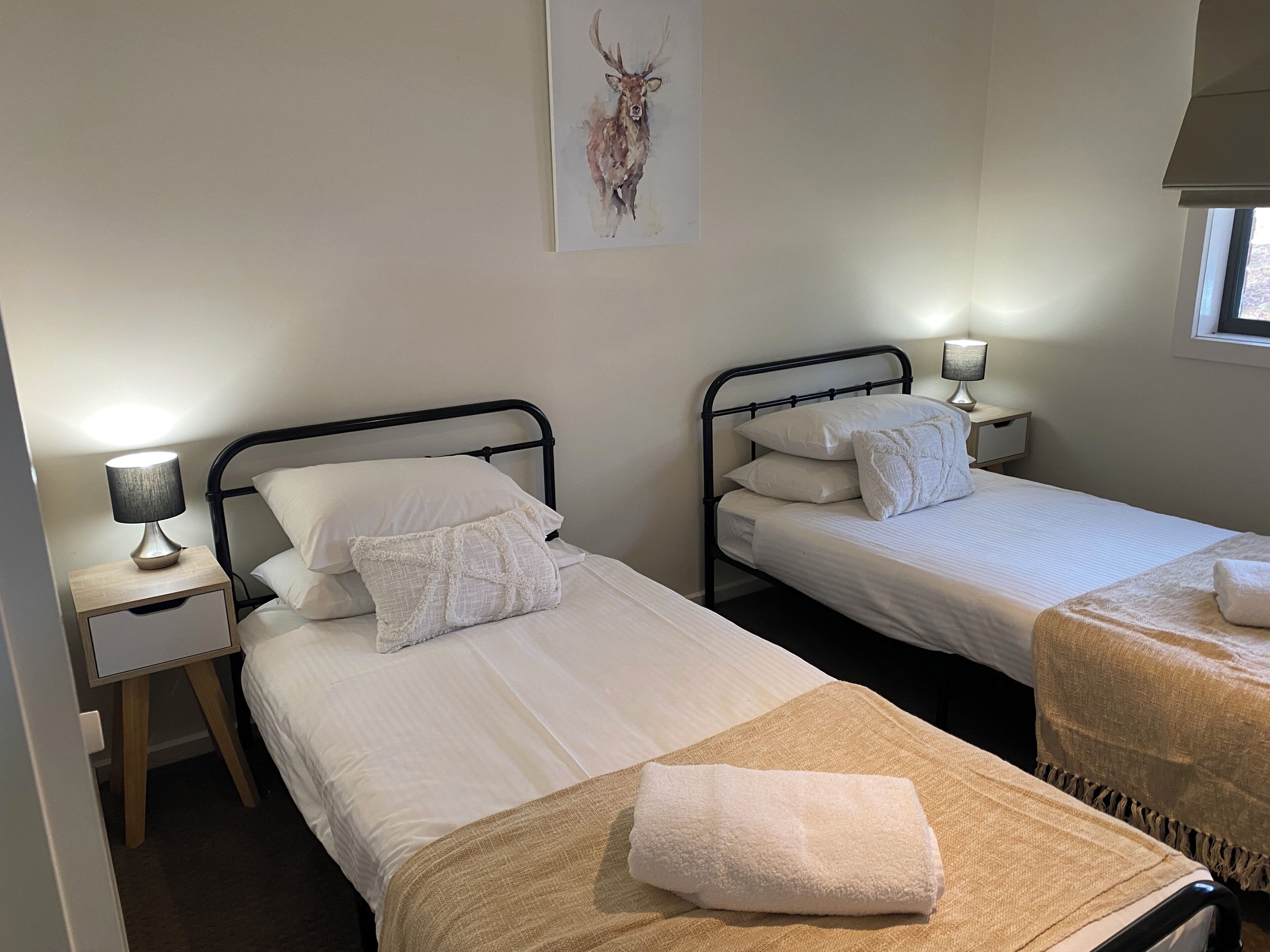 Deux lits simples avec du linge de lit blanc, des tables de chevet avec des lampes, une œuvre d’art encadrée au mur et une fenêtre avec des stores.