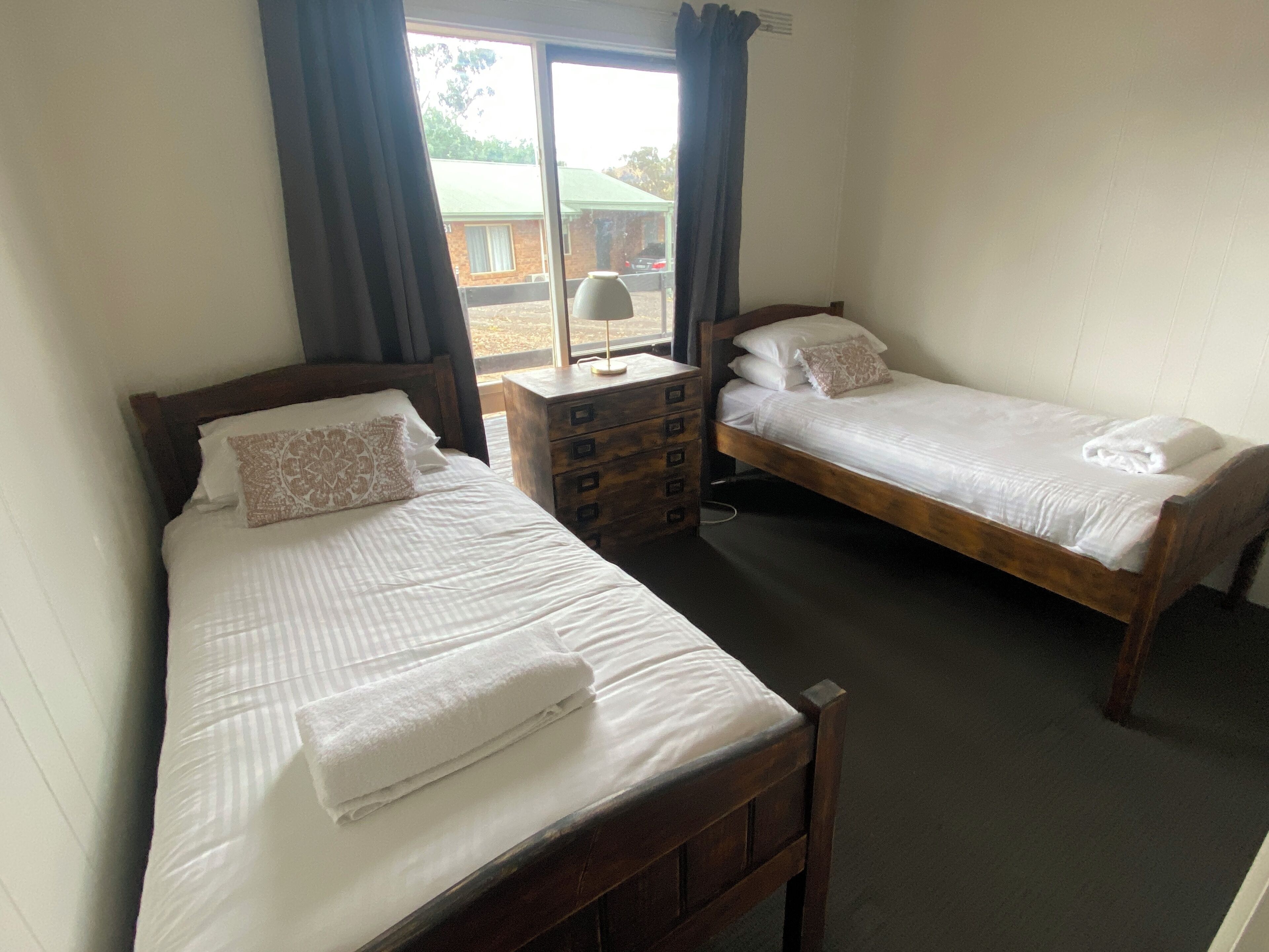 Una habitación con dos camas de madera, cada una con ropa blanca y una almohada. Hay una mesita de noche de madera con una lámpara entre las camas.