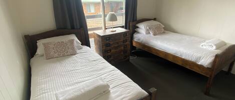 Una habitación con dos camas de madera, cada una con ropa blanca y una almohada. Hay una mesita de noche de madera con una lámpara entre las camas.
