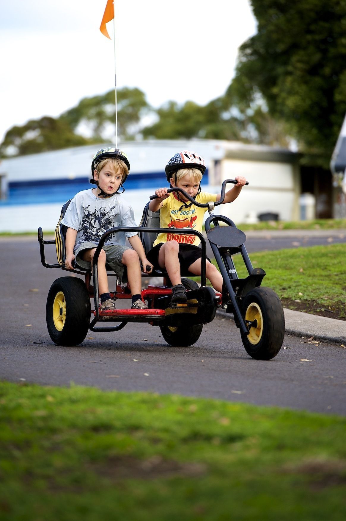 Two children riding a three-wheeled vehicle on a paved road.