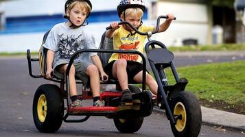 Two children riding a three-wheeled vehicle on a paved road.