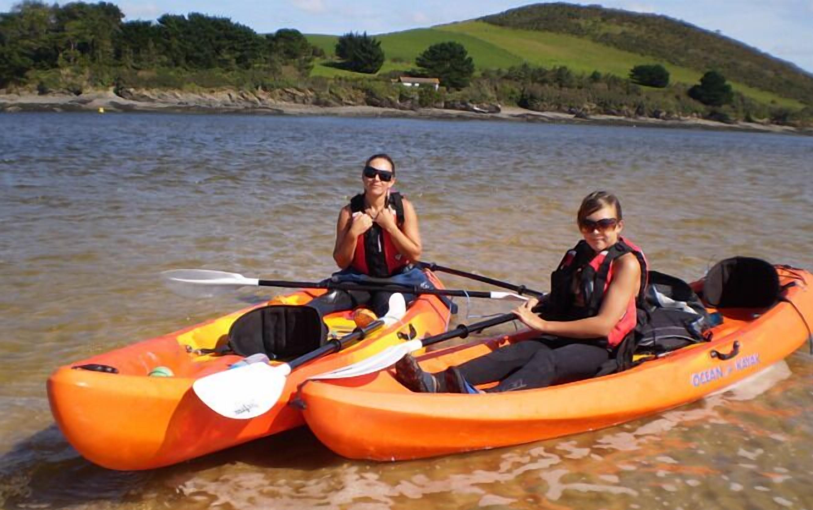 Dos personas en kayak naranja en un lago tranquilo, con una casa al fondo.
