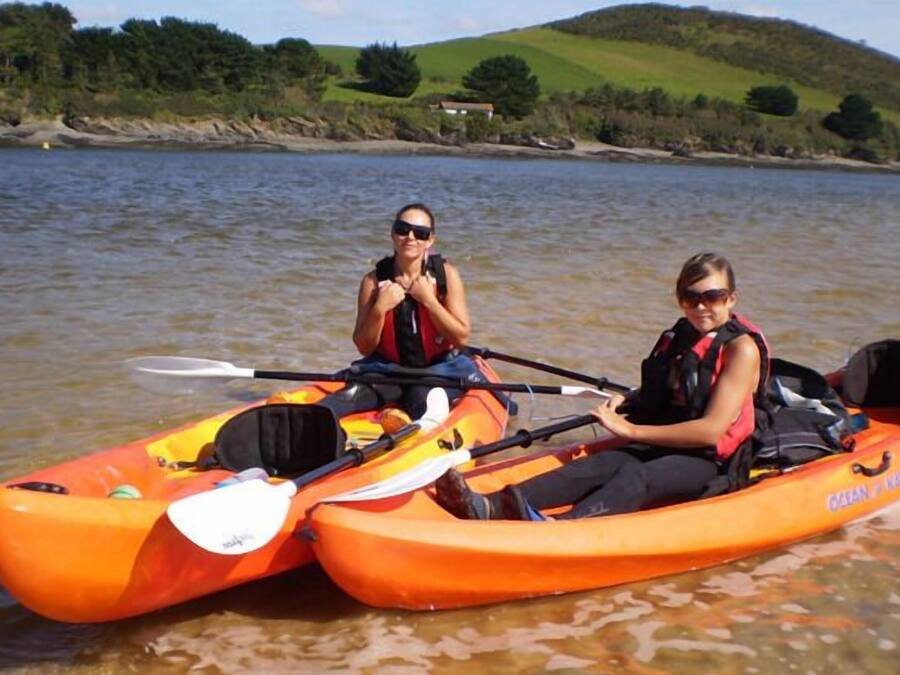 Dos personas en kayak naranja en un lago tranquilo, con una casa al fondo.
