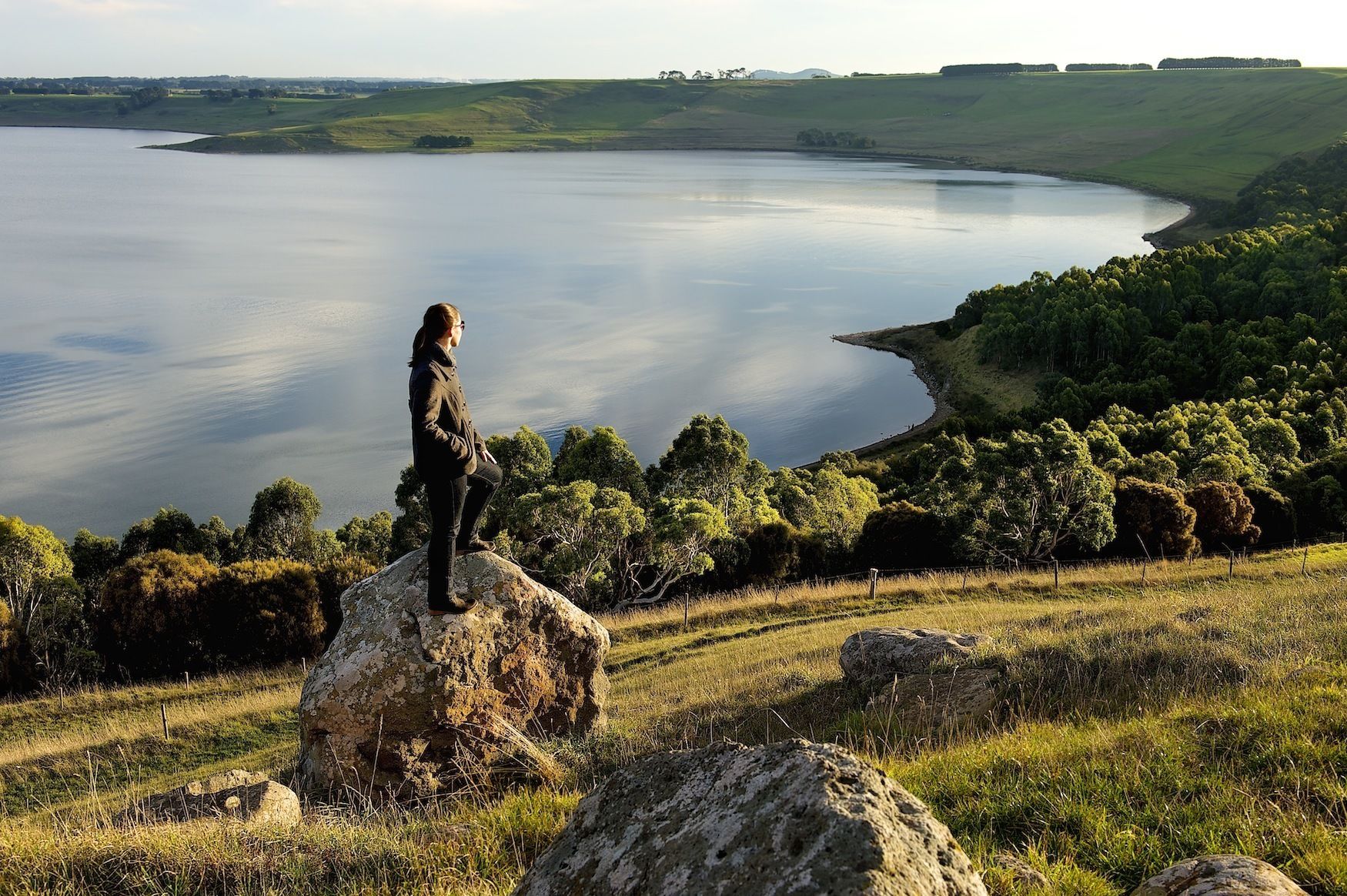 Una persona de pie sobre una roca, contemplando un lago tranquilo con un paisaje boscoso al fondo.