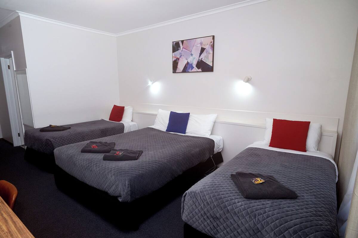 Quarto de hotel com três camas, cada uma com colcha acolchoada, almofada vermelha e branca e um pequeno objeto sobre a cama. Há uma pintura na parede e uma porta à esquerda.