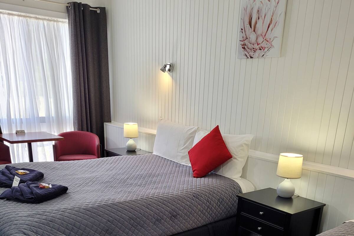 Quarto de hotel com duas camas, uma escrivaninha, uma cadeira e uma janela com cortinas.