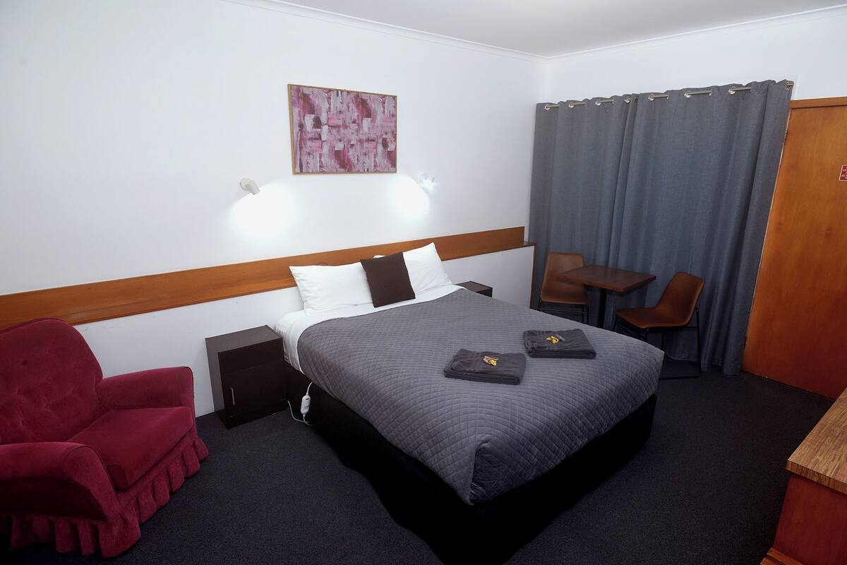 Quarto de hotel com cama, poltrona vermelha, mesa pequena e escrivaninha.