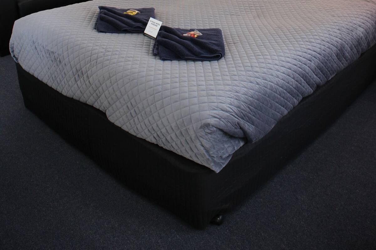 Cama bem arrumada com colcha cinza acolchoada, travesseiro azul escuro e uma toalha dobrada por cima. Um sofá preto com almofada listrada está à esquerda. Um quadro abstrato fixado na parede está acima da cama e uma mesa pequena com abajur ao lado da cama.
