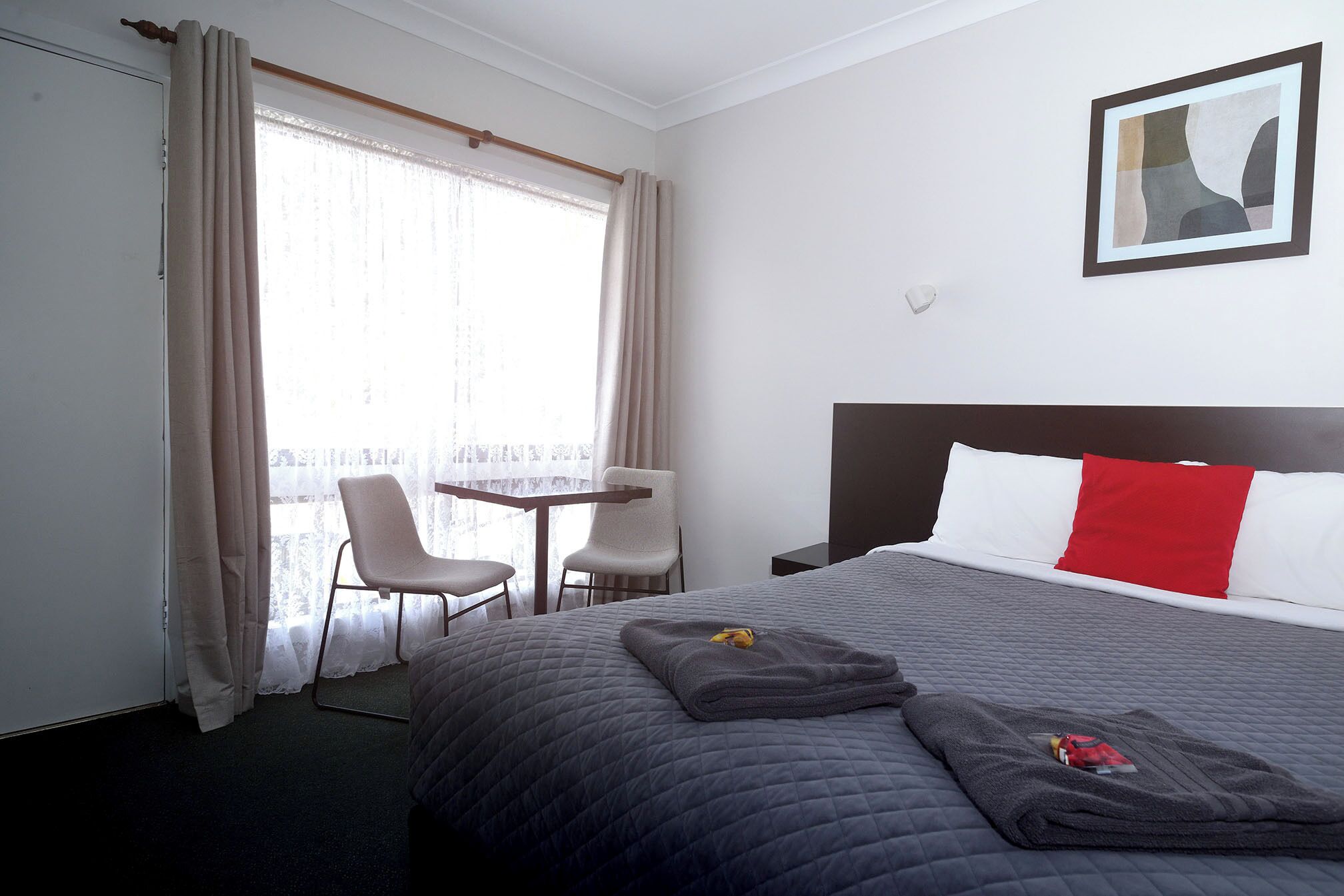 A hotel room with a bed, a desk, two chairs, a window with curtains, and a painting on the wall.