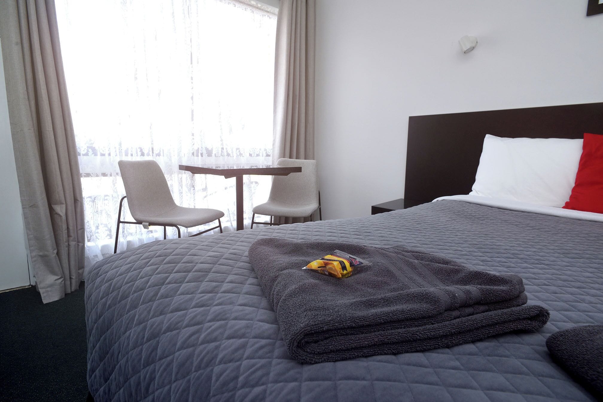 A hotel room with a bed, a small table, two chairs, and a window with curtains.