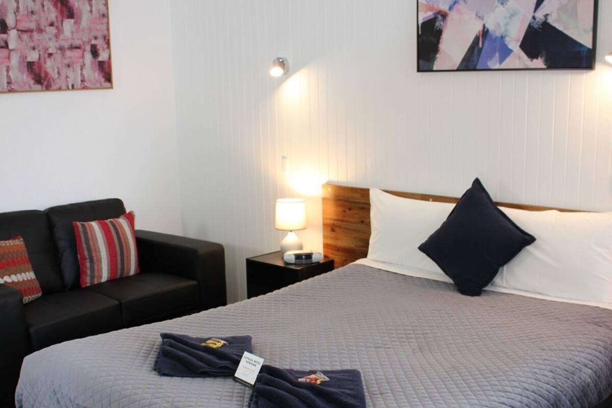 Quarto de hotel com cama, mesa de cabeceira, luminária, sofá e duas pinturas abstratas nas paredes.
