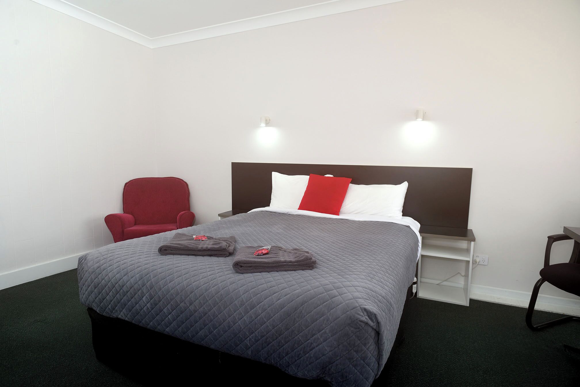 A hotel room with a bed, a red chair, a nightstand, and a wall-mounted light fixture.