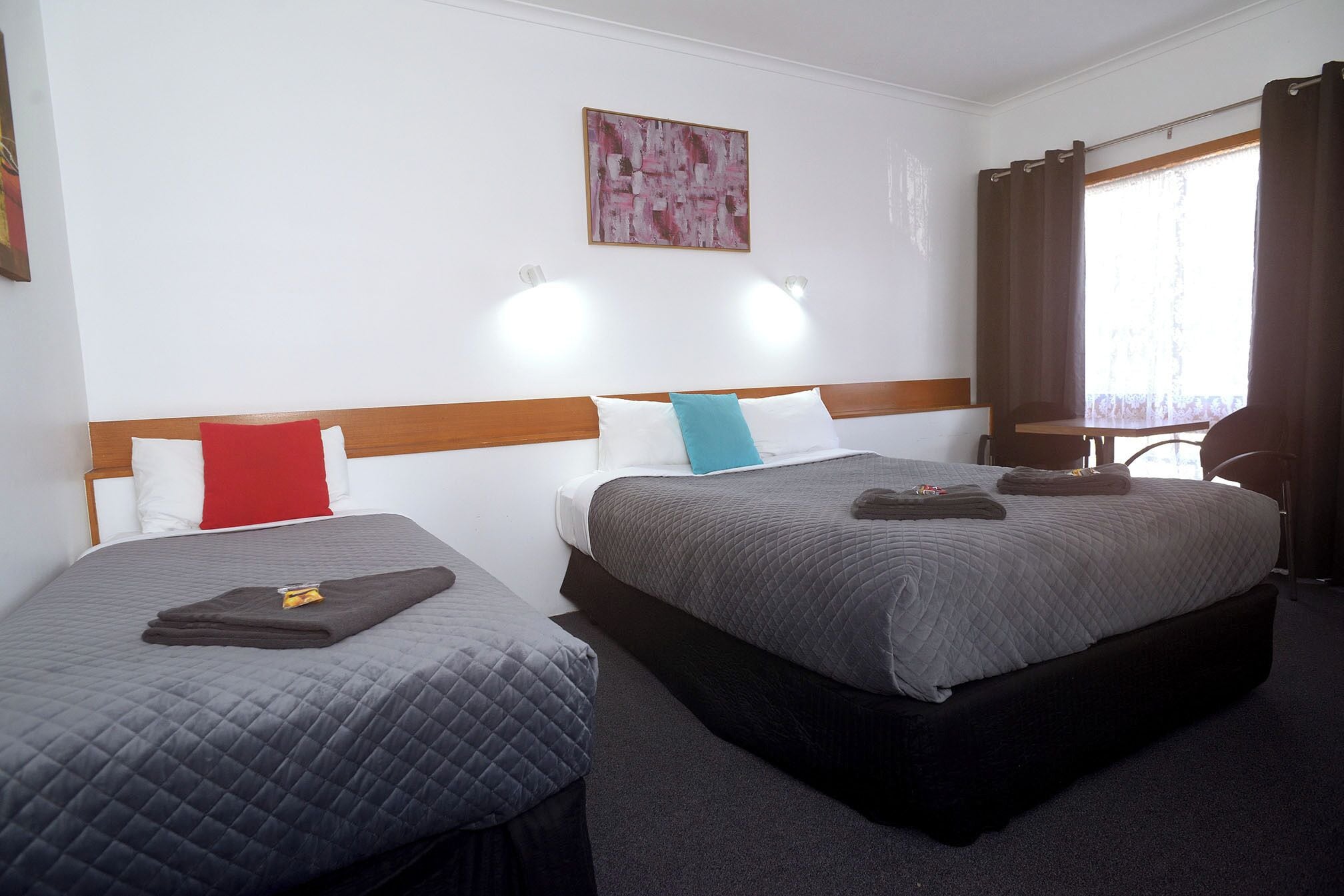 Zwei Einzelbetten mit Kopfteilen, ein kleiner Tisch, ein Stuhl, ein Fenster mit Vorhängen und ein Bild an der Wand.