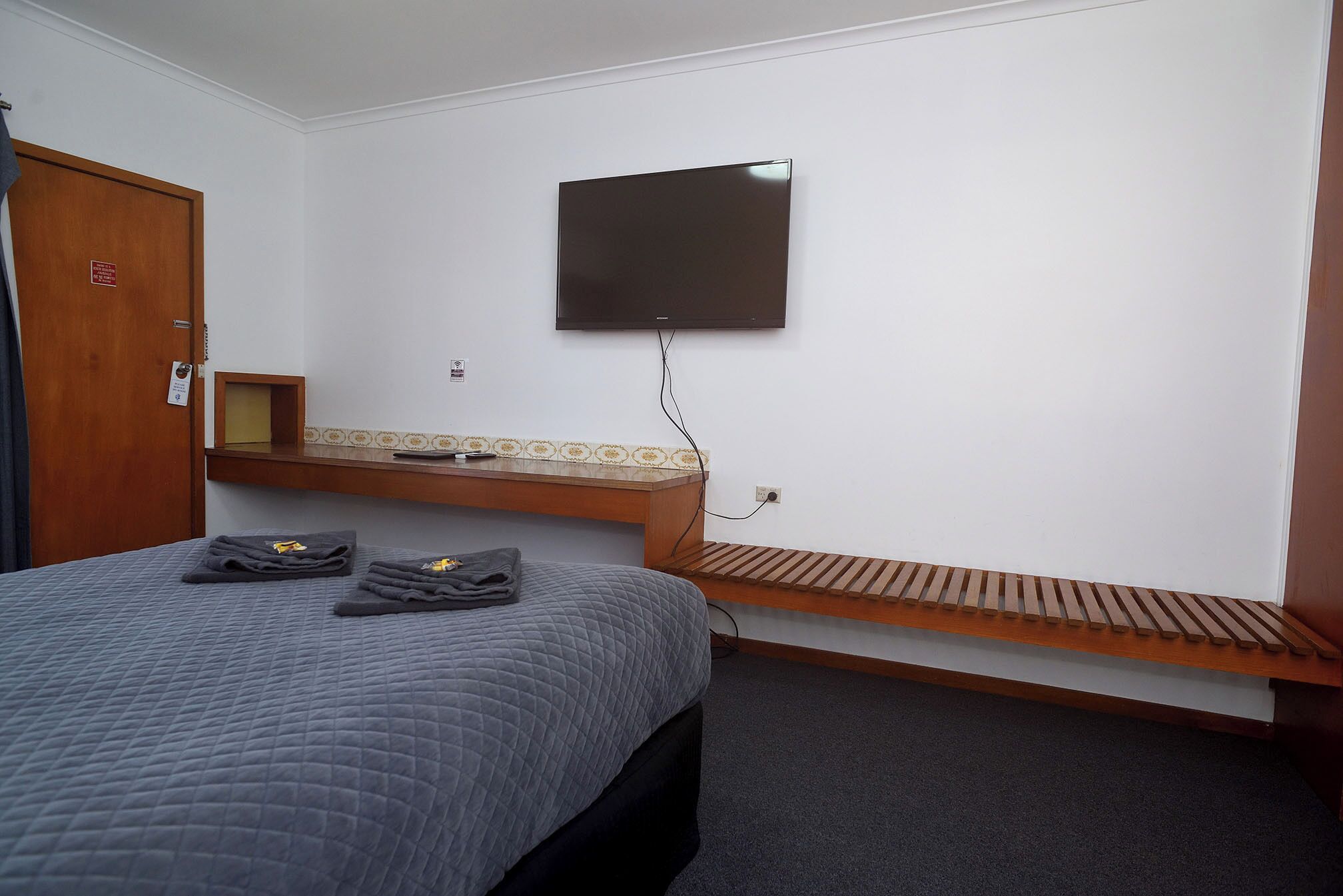 Quarto de hotel com uma cama, uma TV de tela plana na parede, um banco de madeira e uma pequena mesa de madeira.