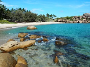 Une plage avec une eau turquoise et limpide, de gros rochers au premier plan et une plage de sable bordée de palmiers à l’arrière-plan.