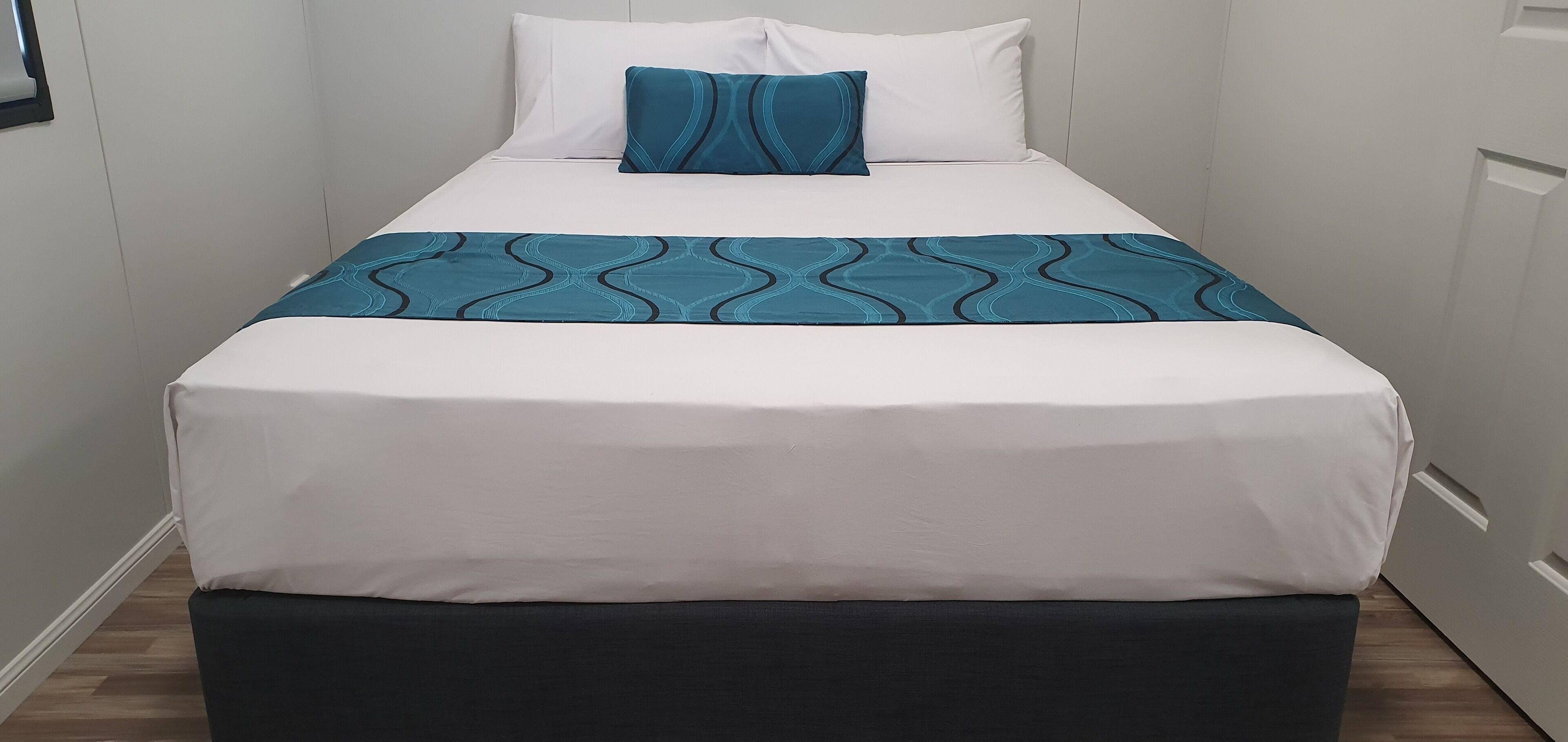 Een net opgemaakt bed met een wit en turquoise beddekleed, een turquoise kussen en een zwart bankje aan het voeteneinde.
